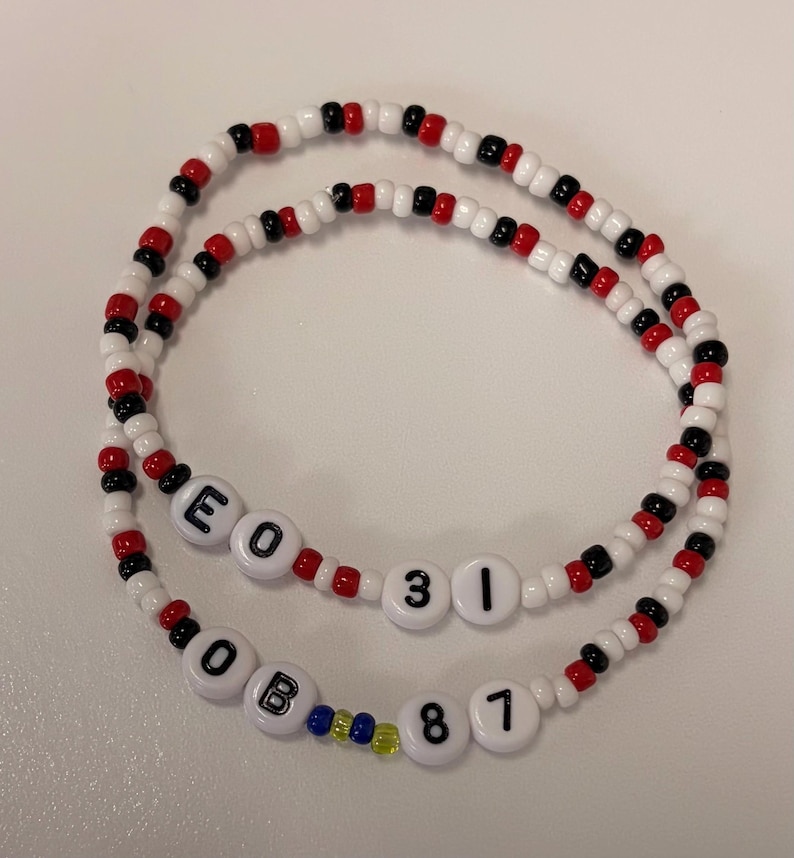 May include: Two beaded bracelets featuring a repeating pattern of white, red, and black seed beads. One bracelet includes white letter and number beads spelling E O 3 1, while the other features white beads spelling O B 8 7 with small blue and yellow accent beads.