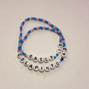 May include: Two beaded bracelets made of small blue, pink, and white seed beads. One bracelet features white letter beads spelling GASLY and the number 10. The second bracelet features white letter beads spelling COLAPINTO and the number 43.
