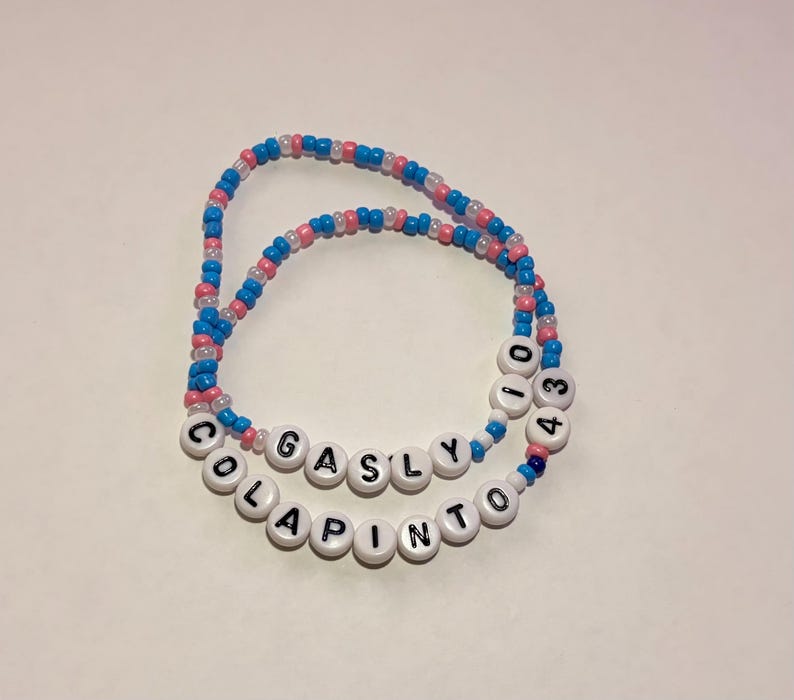May include: Two beaded bracelets made of small blue, pink, and white seed beads. One bracelet features white letter beads spelling GASLY and the number 10. The second bracelet features white letter beads spelling COLAPINTO and the number 43.
