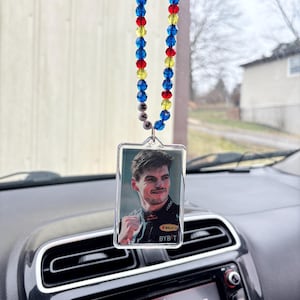 May include: A car air freshener featuring a portrait of a man in a racing uniform. The air freshener is rectangular and hangs from a beaded strand with blue, red, and yellow beads. The car's dashboard and radio are visible in the foreground.