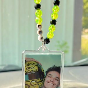 Peut inclure: Un accessoire de voiture avec une photo d'une personne souriante tenant un casque de course jaune et noir. Le charme est suspendu à une chaîne de perles vertes, noires et blanches. Les lettres "L N 4" sont épelées sur les perles.