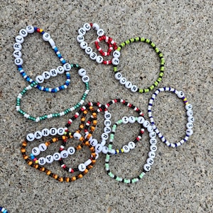 May include: A collection of circular beaded bracelets featuring white letter beads spelling names like Carlos, Lance, Leclerc, Nico, Lando, Oscar, Bear, and Antonelli, interspersed with colorful plastic beads and numerical beads including 5, 18, 27, 81, 87, and 9.