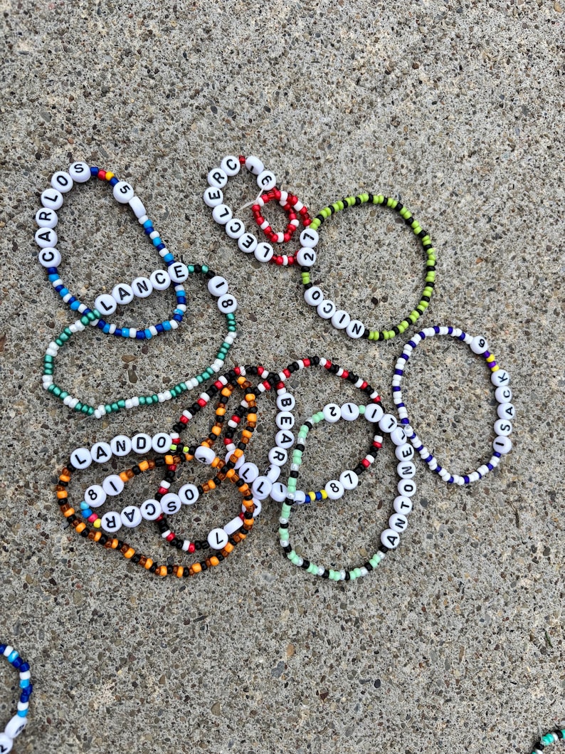 May include: A collection of circular beaded bracelets featuring white letter beads spelling names like Carlos, Lance, Leclerc, Nico, Lando, Oscar, Bear, and Antonelli, interspersed with colorful plastic beads and numerical beads including 5, 18, 27, 81, 87, and 9.