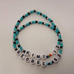 May include: Two circular beaded bracelets featuring turquoise, black, and silver seed beads. One bracelet displays the white letter beads spelling KIMI 12, while the other features white letter beads spelling GEORGE 63, with small multi-colored accent beads.