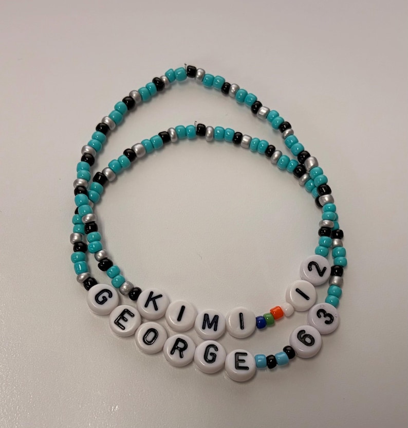 May include: Two circular beaded bracelets featuring turquoise, black, and silver seed beads. One bracelet displays the white letter beads spelling KIMI 12, while the other features white letter beads spelling GEORGE 63, with small multi-colored accent beads.