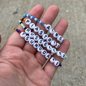 May include: Four beaded bracelets featuring white letter beads spelling out PIASTRI 81, ALBON 23, RUSSELL 63, and TSUNODA 22. Each bracelet is constructed with small, colorful plastic beads in various patterns, including blue, white, green, red, and yellow.