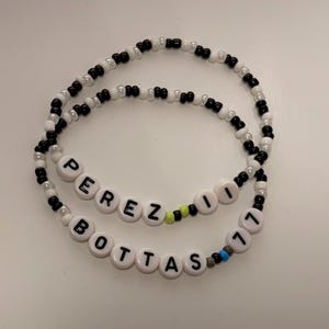 May include: Two beaded bracelets made of small black and white beads. One bracelet features white letter beads spelling PEREZ 11 with a lime green bead. The other bracelet features white letter beads spelling BOTTAS 77 with a blue and grey bead.
