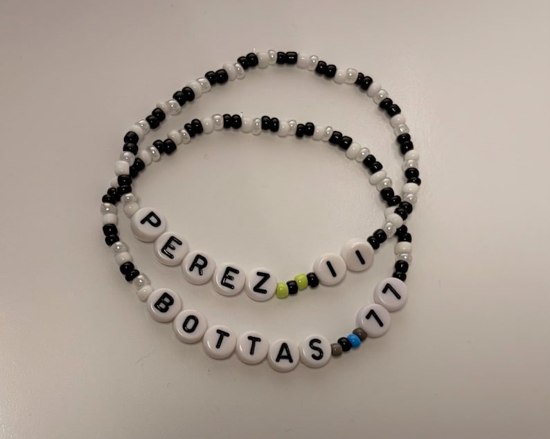 May include: Two beaded bracelets made of small black and white beads. One bracelet features white letter beads spelling PEREZ 11 with a lime green bead. The other bracelet features white letter beads spelling BOTTAS 77 with a blue and grey bead.