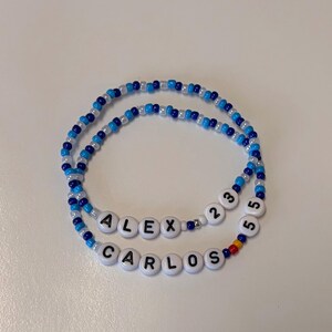 May include: Two beaded bracelets featuring a mix of blue, light blue, and white seed beads. One bracelet displays the name ALEX followed by the numbers 23, and the other displays the name CARLOS followed by the number 55 in white letter beads.