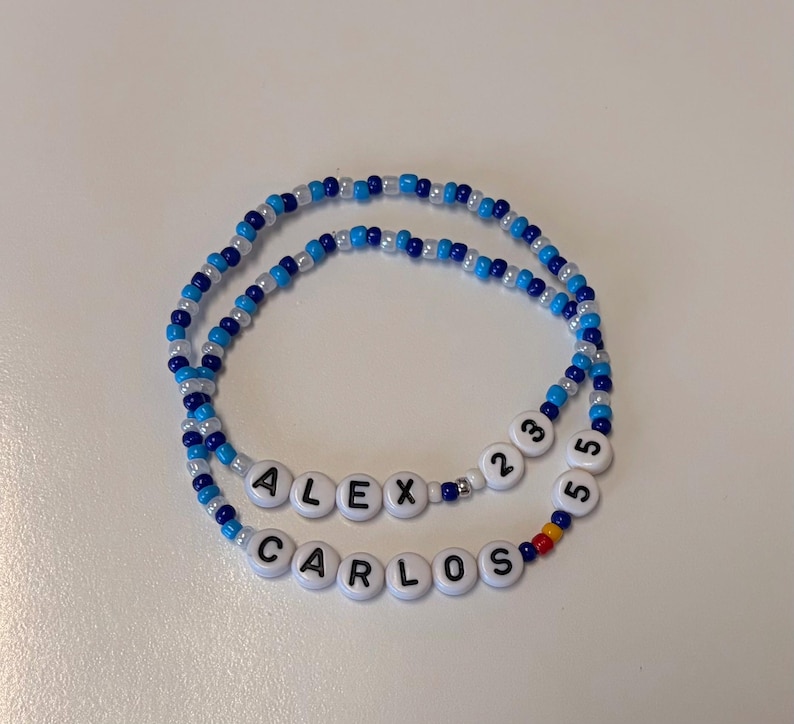 May include: Two beaded bracelets featuring a mix of blue, light blue, and white seed beads. One bracelet displays the name ALEX followed by the numbers 23, and the other displays the name CARLOS followed by the number 55 in white letter beads.