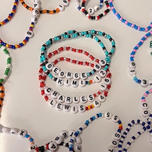 Peut inclure: Plusieurs bracelets de perles avec des perles de lettres blanches épelant des noms comme "GEORGE" et "CHARLES". Les bracelets sont de différentes couleurs, dont le rouge, le bleu et le turquoise, avec des perles de chiffres.
