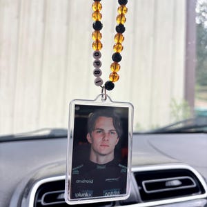 May include: Clear rectangular car charm featuring a portrait of a young man in a black racing suit. The charm hangs from a beaded strand with black and amber beads. The word "OP81" is spelled out in white beads.