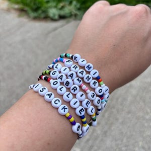 May include: A stack of five beaded bracelets featuring white round beads with black letters spelling out the names KIMI, FRANK, LIAM, GABRIEL, and ISACK. The bracelets are constructed with colorful small seed beads in various patterns and color combinations.