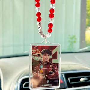 May include: Clear acrylic car charm featuring a portrait of a race car driver in a red racing suit. The charm hangs from a beaded strand of red and clear beads. The text "CL6" is visible on the strand.