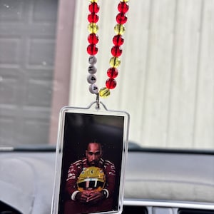 May include: A car air freshener featuring a photo of a person in a racing suit holding a helmet. The air freshener is suspended by a beaded chain with red, yellow, and white beads. The letters "LHA" and the number "4" are spelled out on the chain.