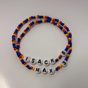 May include: Two circular beaded bracelets featuring a repeating pattern of small blue, yellow, and red glass beads. One bracelet includes white round letter beads spelling ISACK and the number 6. The other bracelet features white letter beads spelling MAX and the number 3.
