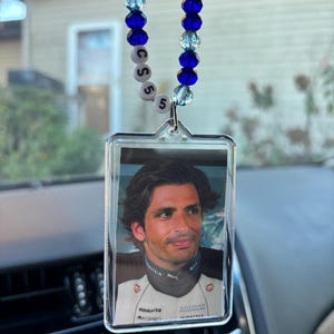 May include: A car charm featuring a photograph of a man in a racing uniform. The charm is encased in a clear, rectangular frame and hangs from a beaded strand of blue and clear beads. The charm has the letters "CSS" and the number "5" on it.