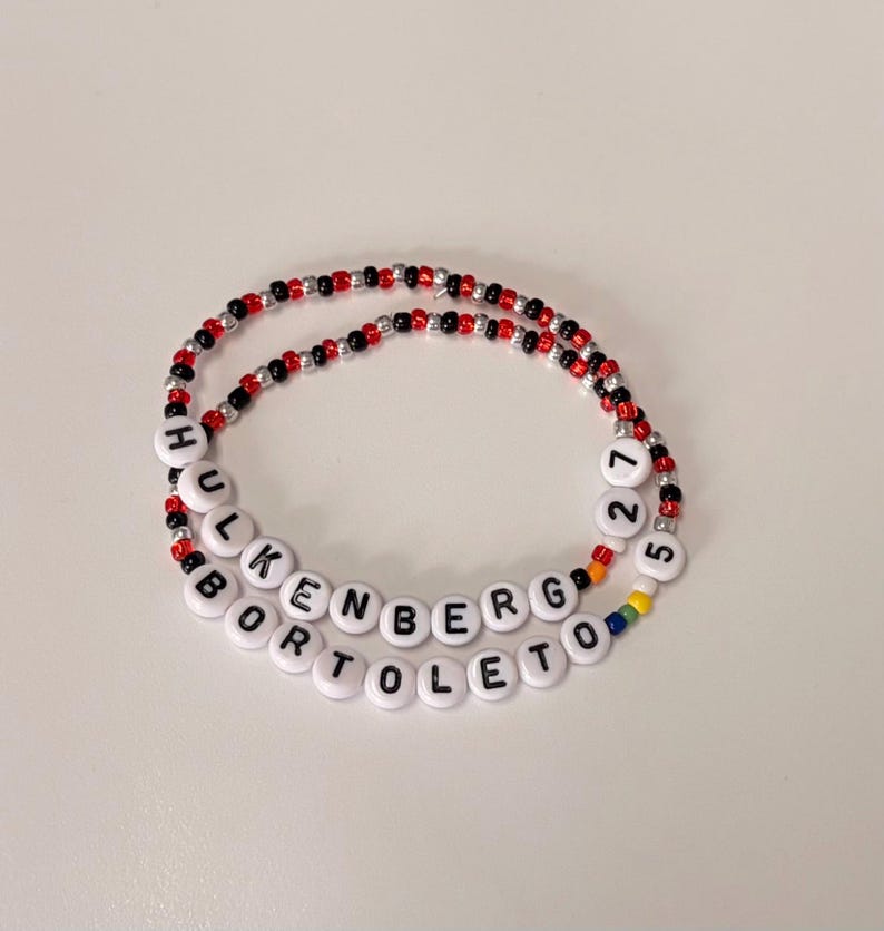 May include: Two beaded bracelets featuring alternating red, black, and silver metallic beads. One bracelet displays the white letter beads spelling HULKENBERG 27, while the other features white letter beads spelling BORTOLETO 5 with small rainbow-colored beads.