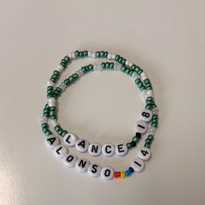 May include: Two beaded bracelets featuring small green and white seed beads. One bracelet displays the text LANCE 18 in white letter beads. The other bracelet features the text ALONSO 14 with small red, yellow, and blue accent beads.