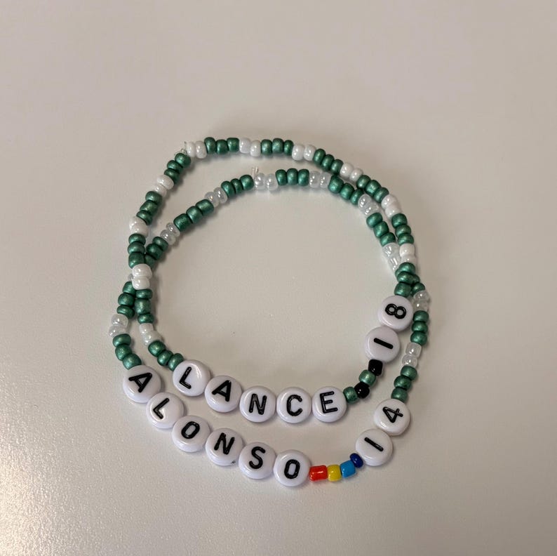 May include: Two beaded bracelets featuring small green and white seed beads. One bracelet displays the text LANCE 18 in white letter beads. The other bracelet features the text ALONSO 14 with small red, yellow, and blue accent beads.