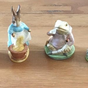 Può includere: Una collezione di statuine in ceramica raffiguranti personaggi di una storia per bambini. Le statuine includono un coniglio avvolto in uno scialle rosso, un coniglio con un vestito blu, una rana e due conigli che leggono un libro. Le statuine sono dipinte con colori pastello tenui.