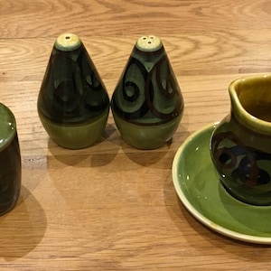 Może przedstawiać: Ceramiczny zestaw kuchenny w kolorze zielono-brązowym. Zestaw zawiera słoik z pokrywką, dwie solniczki i pieprzniczki oraz mały dzbanuszek do śmietanki z pasującym spodkiem. Elementy mają błyszczące wykończenie i dekoracyjne wzory.