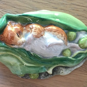 以下が含まれることがあります： 緑色のエンドウ豆のさやの中で眠っているハムスターの陶器製の置物。 ハムスターは茶色と白の毛皮で、エンドウ豆のさやは光沢のある仕上げの鮮やかな緑色です。 置物は木製の表面にあります。