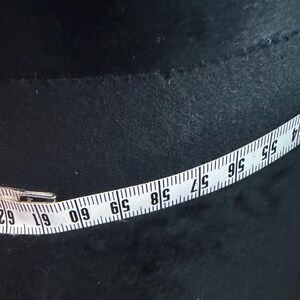 May include: A white measuring tape with black numbers and markings is laid across a dark, textured surface. The tape shows measurements from 55 to 62 inches. The background is a dark, possibly black, material.