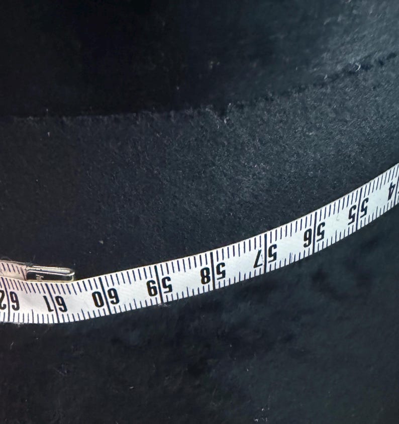 May include: A white measuring tape with black numbers and markings is laid across a dark, textured surface. The tape shows measurements from 55 to 62 inches. The background is a dark, possibly black, material.