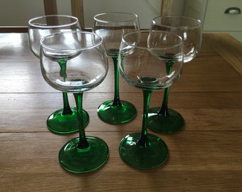 May include: Six clear glass wine glasses with green stems and bases. The glasses are arranged in a semi-circle on a wooden surface.