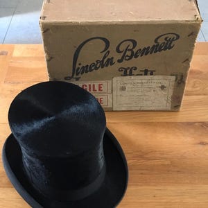 May include: A black top hat with a black band sits in front of a cardboard box. The box is labeled "Lincoln Bennett" in a vintage font. The hat has a tall, cylindrical crown and a wide brim. The box has shipping labels.