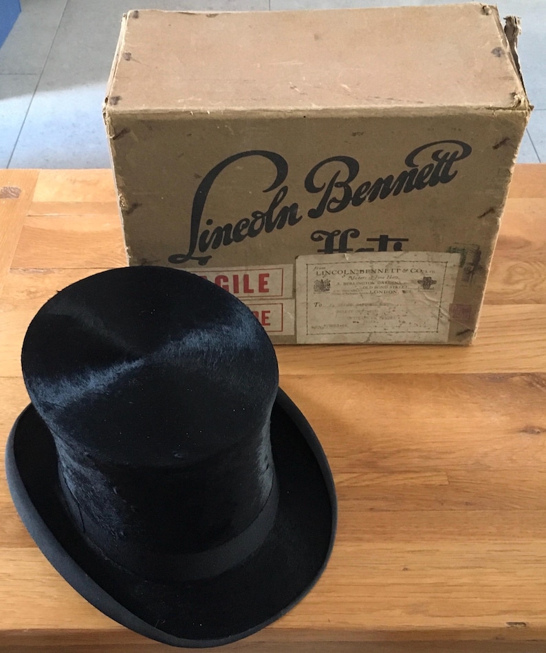 May include: A black top hat with a black band sits in front of a cardboard box. The box is labeled "Lincoln Bennett" in a vintage font. The hat has a tall, cylindrical crown and a wide brim. The box has shipping labels.