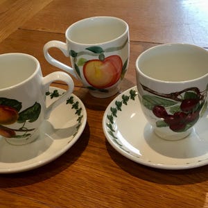 Può includere: Tre tazze da tè e piattini in ceramica bianca con disegni di frutta. Una tazza presenta una pesca, un'altra ciliegie e la terza una mela. I piattini hanno un bordo di foglie verdi. Le tazze e i piattini sono su una superficie di legno.
