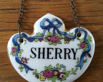 May include: A white ceramic Sherry label with a heart shape, featuring a blue ribbon and floral design. The word "SHERRY" is in black capital letters. The label is suspended by a delicate chain.