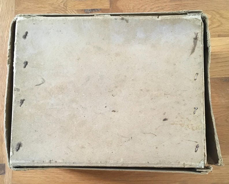 May include: An open, rectangular cardboard box with a light brown lid. The lid is secured with small metal staples. The box is sitting on a wooden surface.