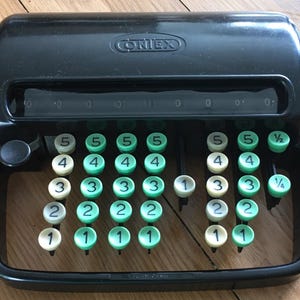 Op de afbeelding: Een vintage zwarte Contex-rekenmachine met witte en groene cijfertoetsen. De rekenmachine heeft een reeks cijfertoetsen, inclusief breuken. De bovenkant van de rekenmachine heeft een displayvenster en de merknaam "CONEX".