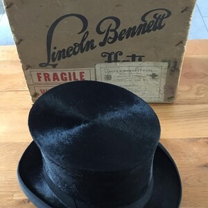 May include: A black top hat with a rounded crown and a wide brim, sitting in front of a cardboard box. The box has the words "Lincoln Bennett" printed on it. The hat is a classic formal accessory.