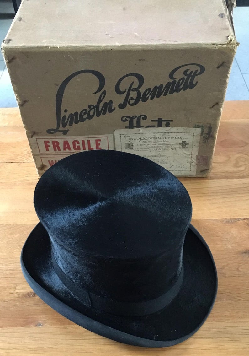 May include: A black top hat with a rounded crown and a wide brim, sitting in front of a cardboard box. The box has the words "Lincoln Bennett" printed on it. The hat is a classic formal accessory.