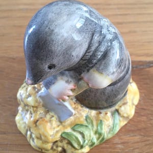 May include: A ceramic figurine of a grey mole digging in the ground with a shovel. The mole is holding the shovel with its front paws and has a small, black eye. The ground is yellow with green leaves.