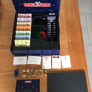 May include: A Waddingtons European Edition Monopoly board game set featuring a dark blue box with gold stars, paper money in various denominations, green and red plastic houses and hotels, game cards, gold-colored tokens, two blue dice, and a manual.