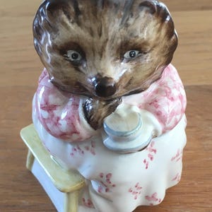 May include: A ceramic figurine of a hedgehog wearing a pink dress and a white apron, sitting on a yellow stool and holding a teacup.