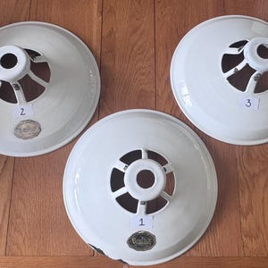 May include: Three white, vintage light shades with a circular design. Each shade features a central opening with a white, cylindrical component and supporting struts. The shades are numbered 1, 2, and 3, and are displayed on a wooden surface.