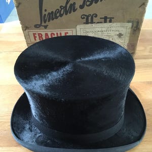 May include: A black top hat with a rounded crown and a wide brim. The hat has a black ribbon around the base of the crown. The hat is sitting on a wooden surface. The box behind the hat has the text "Lincoln Bennett" on it.
