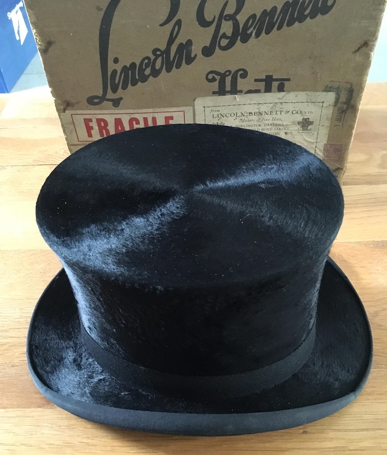 May include: A black top hat with a rounded crown and a wide brim. The hat has a black ribbon around the base of the crown. The hat is sitting on a wooden surface. The box behind the hat has the text "Lincoln Bennett" on it.