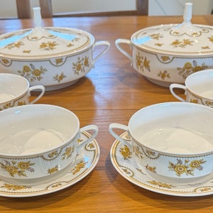 Może przedstawiać: Zestaw sześciu białych porcelanowych misek do zupy z złotymi wzorami kwiatowymi. Miski występują w dwóch rozmiarach, większe miski mają pokrywki. Miski są ułożone na drewnianym stole.