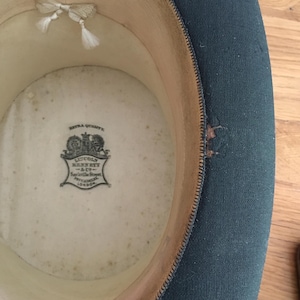 May include: An inside view of a dark green top hat. The interior is cream-colored with a printed emblem and a small white bow. The hat's brim is lined with a light brown material.