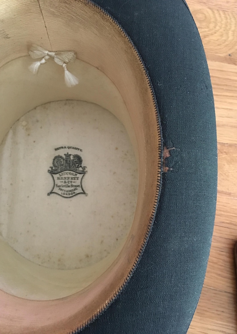 May include: An inside view of a dark green top hat. The interior is cream-colored with a printed emblem and a small white bow. The hat's brim is lined with a light brown material.