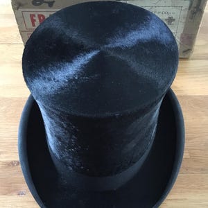 May include: A black top hat with a wide brim and a black band. The hat is made of a soft, velvety material. The hat is sitting on a wooden surface. A cardboard box is in the background.