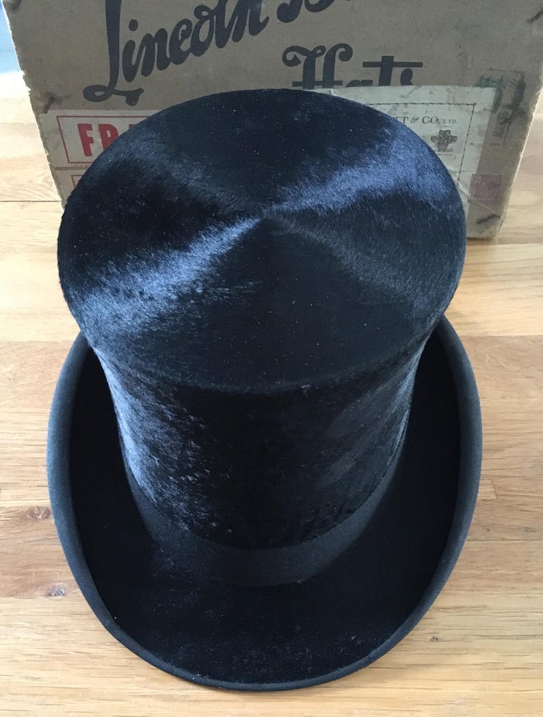 May include: A black top hat with a wide brim and a black band. The hat is made of a soft, velvety material. The hat is sitting on a wooden surface. A cardboard box is in the background.