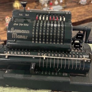 Op de afbeelding: Een vintage Hamann Manus mechanische rekenmachine in donkergroen. De rekenmachine heeft meerdere rijen nummerwielen en een slinger. De tekst "Add. Mult." en "De Te We Hamann Manus" is zichtbaar op de machine.